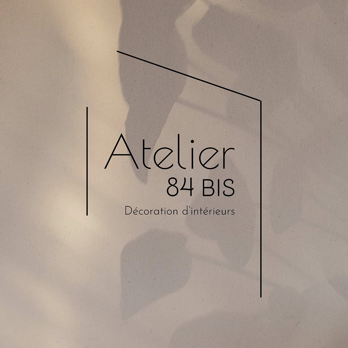 Logo noir de l’Atelier 84 bis avec la mention Décoration d’intérieurs, sur fond beige traversé d’ombres de feuillage