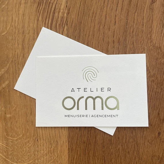 Deux cartes de visite Atelier Orma superposées sur un plateau en chêne, mention « Menuiserie | Agencement »