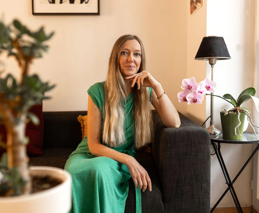 Céline Duval, astrologue, assise sur un canapé gris en robe verte près d’une orchidée, portrait utilisé dans sa communication