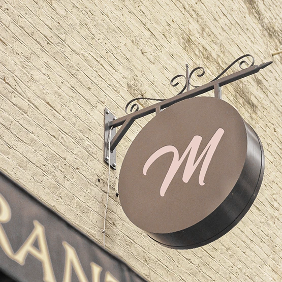Enseigne ronde en potence au monogramme M rose de Chez Marion, mise en situation sur une façade de briques claires