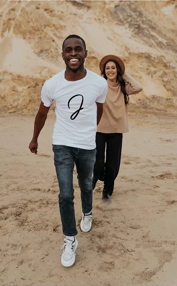T-shirt blanc floqué du J manuscrit de Justine Lorre, porté par un homme souriant sur une plage de sable